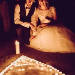 Hochzeitsfeier, Braut und Bräutigam knieend vor Lichtermeer aus Kerzen in herzform outdoor