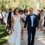 Braut und Bräutigam Hand in Hand laufen auf Kamera zu, Ganzkörper, Hochzeitsstrauß, Verwandtschaft im Hintergrung, outdoor